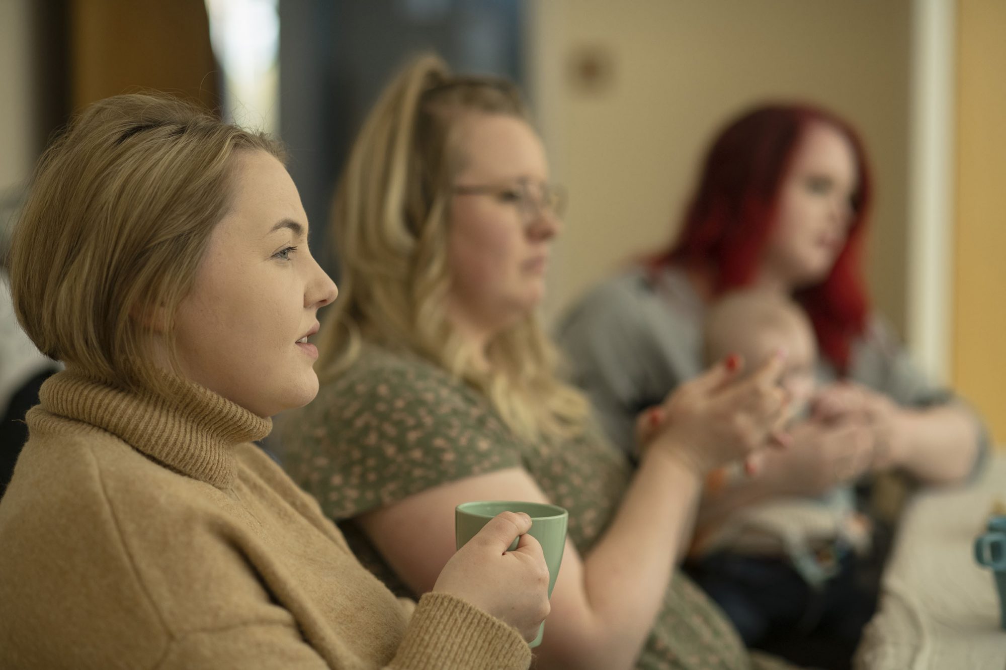 A group of mums sat talking as one holds a cup of tea and another is holding a sleeping baby