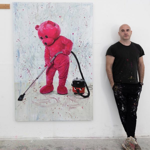 Paul Robinson stood to the right next to a piece of his art, which is an oil painting of his Pink Bear vacuuming with a red Henry vacuum cleaner