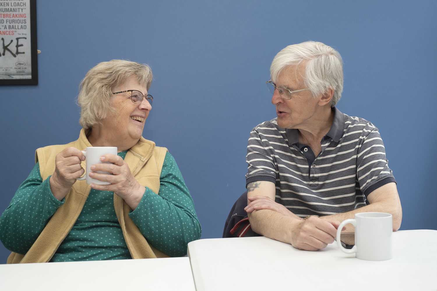 A woman is holding a coffee cup and laughing while talking to a man seated at a table in a room with blue walls