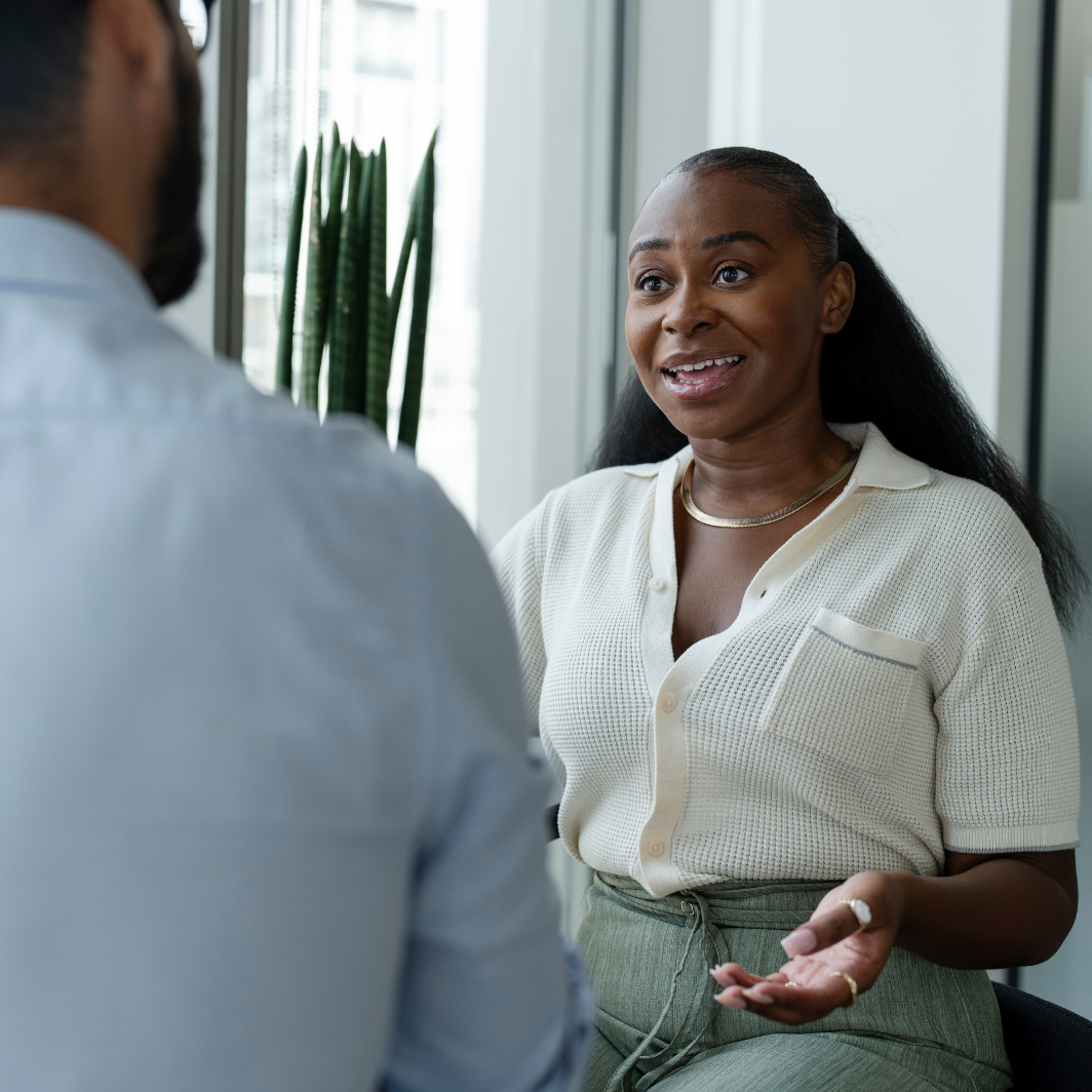 A woman is sitting indoors, speaking to a man in conversation. She looks engaged and expressive, smiling slightly with her hands open as she talks.