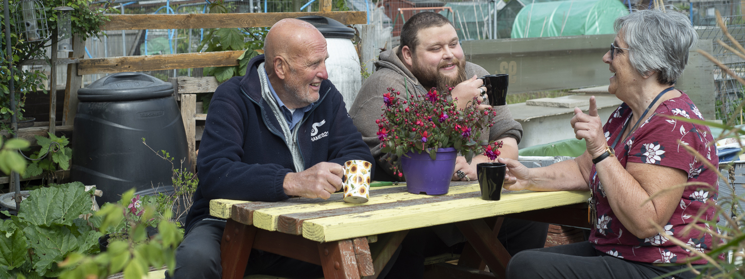 Three people seated at a bench wiith cups of tea at our Open Spaces project allotments in Bexleyheath