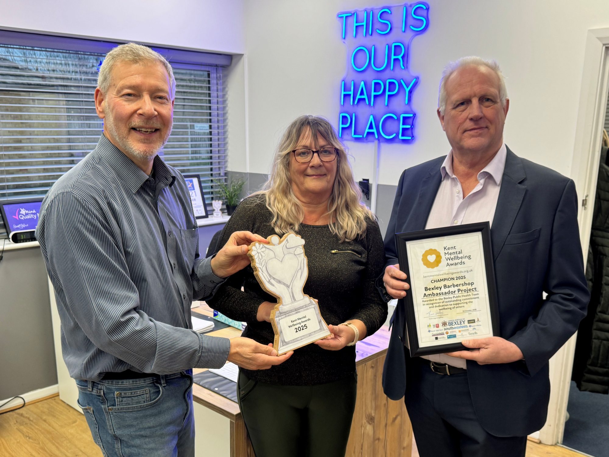 Mind in Bexley Development Lead Simon Dolby is presenting a trophy and certificate to Philippa Wills and Cllr Melvyn Seymour from the London Borough of Bexley. The wooden trophy has Kent Mental Wellbeing Awards engraved on the front and the certificate is in a frame and says Champion 2025 Bexley Barbershop Ambassador Project