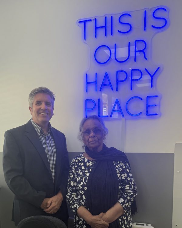 Dr David Palmer and Chair Shaheen Westcombe stood facing the camera next to a neon sign that reads 'THIS IS OUR HAPPY PLACE'
