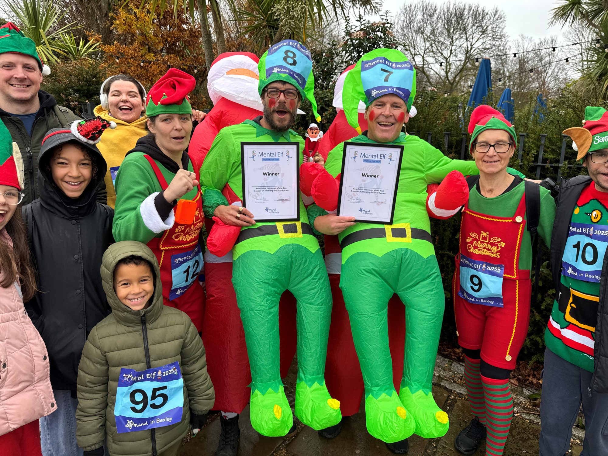 David Wilkins and Steve Edwards are stood holding their Best Dressed Adult Elf certificates in front of a cheering group of young and adult eleves