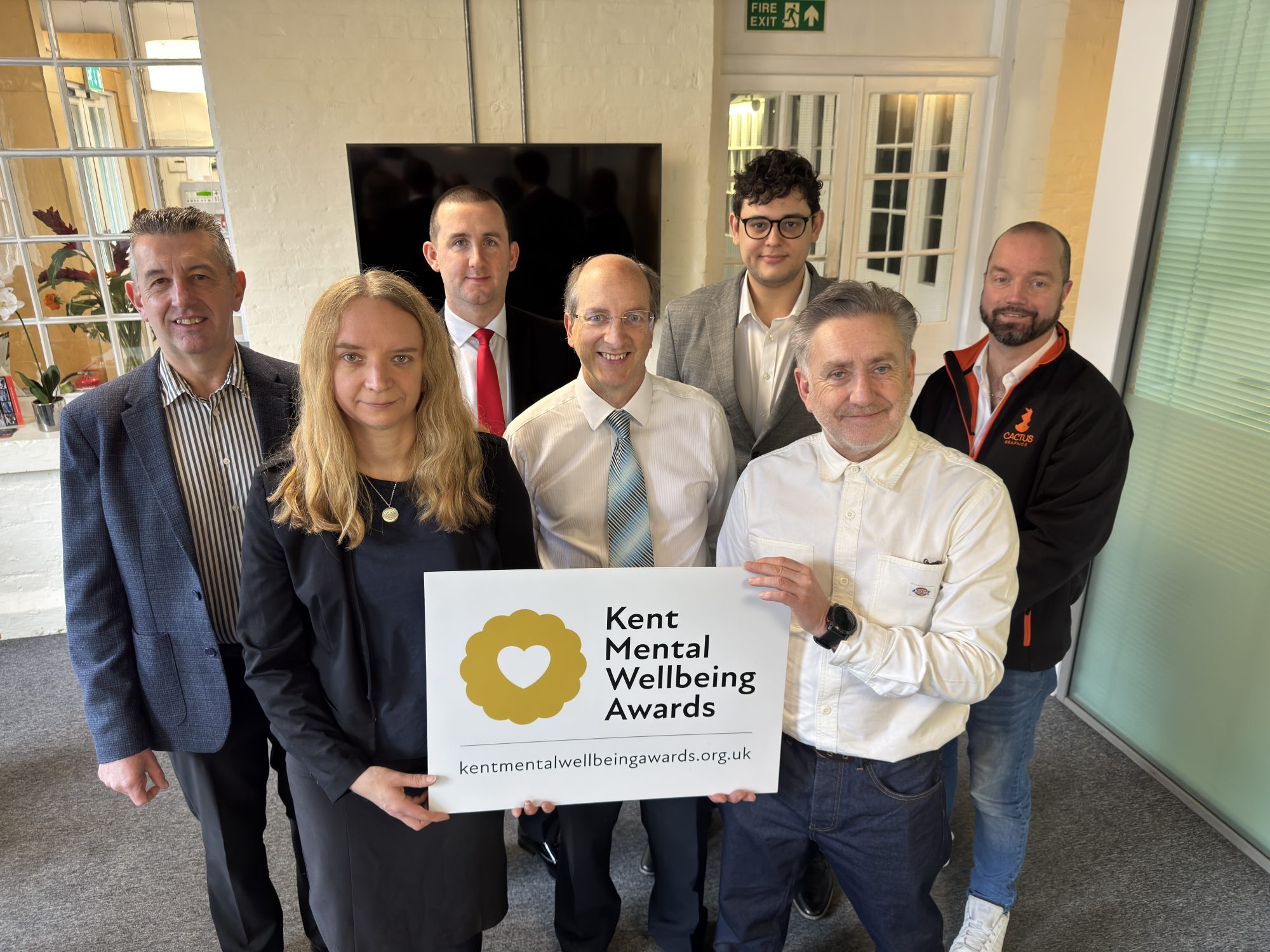 Seven people are stood in a room, with two at the front holding a Kent Mental Wellbeing Awards banner