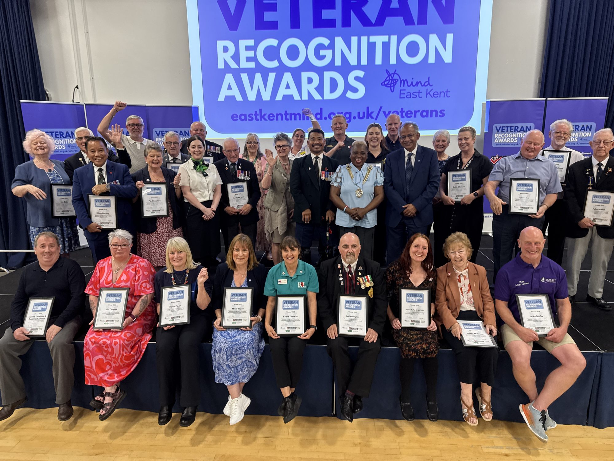 A group of 40 plus veterans hold up their award certificates and smile for the camera at the 2025 ceremony, in front of a large Veterans Recognition Awards banner