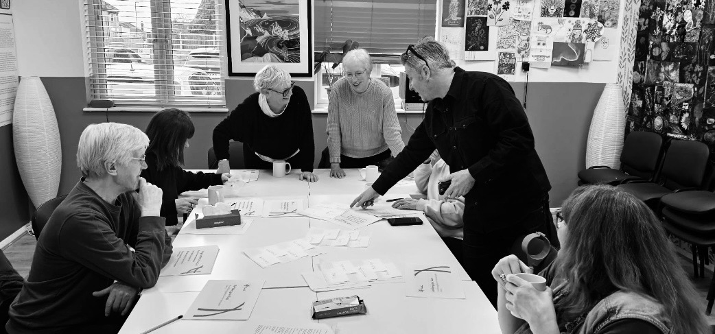 Three members of the carers group are stood discussing their poem, with one pointing at a paper. Four more are seated at a table with one holding a mug