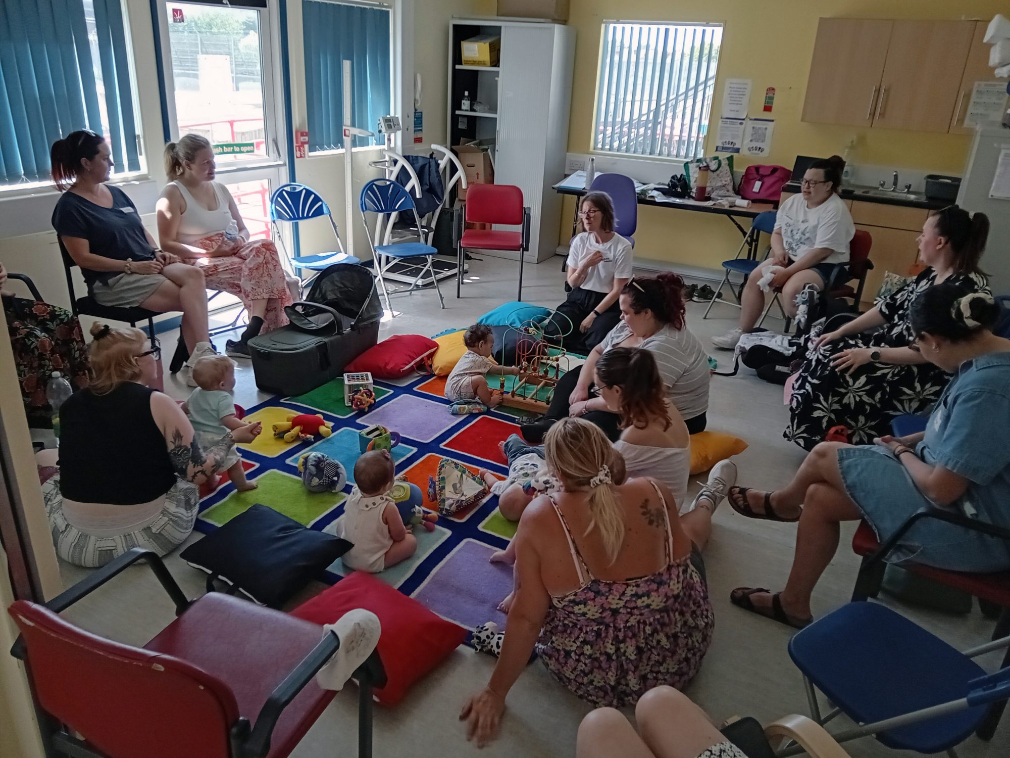 A large group of mums and babies are at the singing session. Some mums are sat on chairs - others on mats wit h their babies
