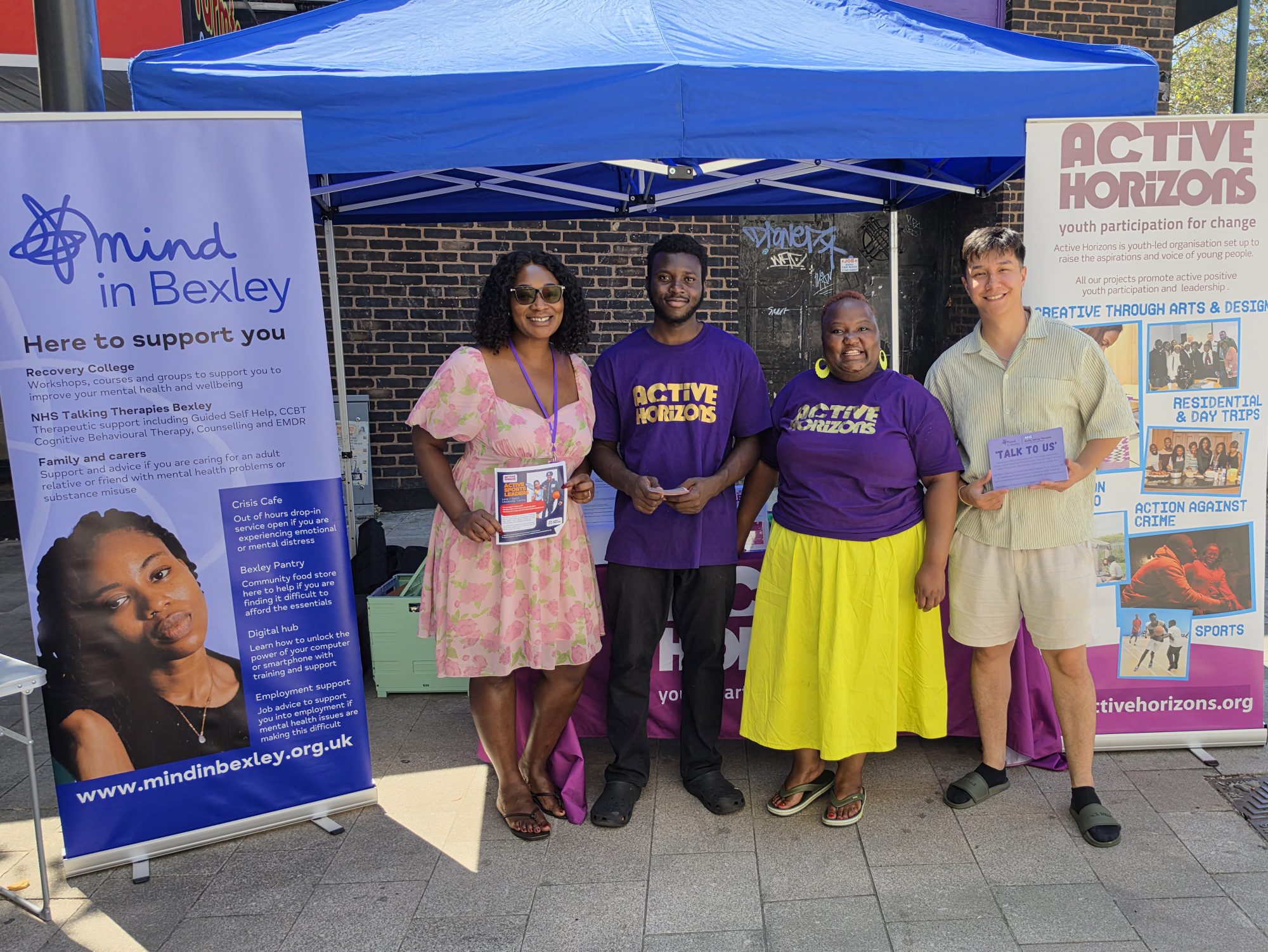 Four people are stood outside smiling on a hot and sunny day. There are Mind in Bexley and Active Horizons roller banners and promotional materials
