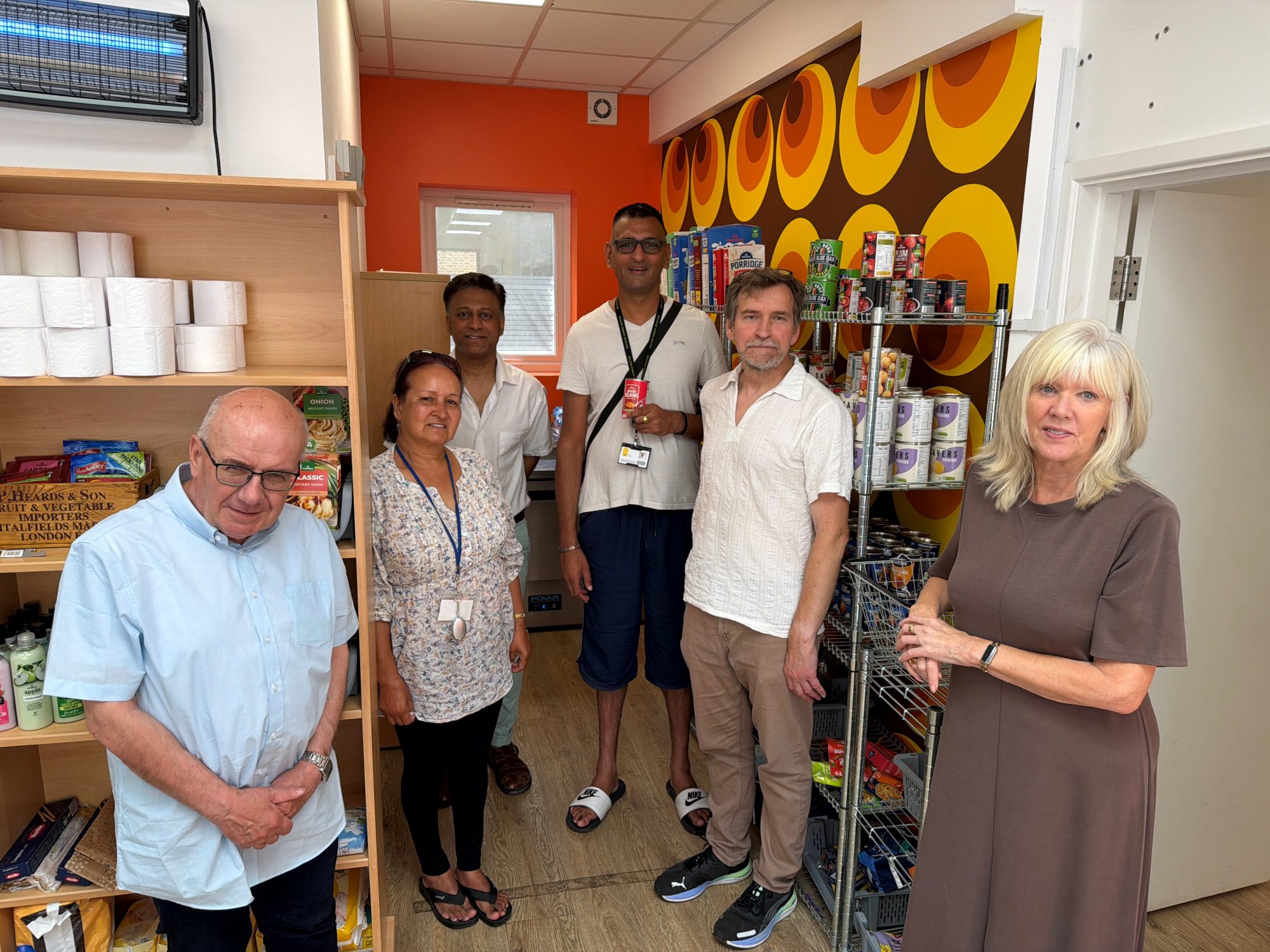 Six people stand inside the Bexley Community Pantry, surrounded by shelves stocked with tinned food, cereal, toiletries and other donated essentials.